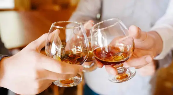 Manos brindando con copas de brandy ámbar. ¿Qué es el brandy? Es un espíritu destilado de vino, perfecto para celebrar.