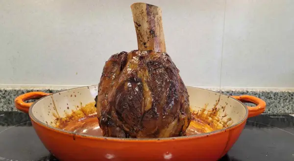 Un jugoso jarrete de ternera asado, con su hueso expuesto, reposa en una cazuela naranja, listo para ser disfrutado.