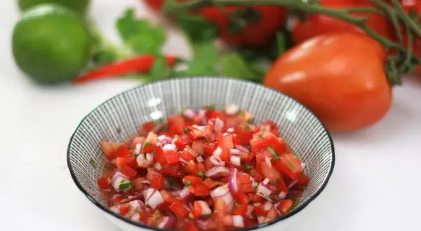 Un tazón de pico de gallo fresco, con tomates, cebolla morada y cilantro picados. Al fondo, tomates, limas y chiles rojos.