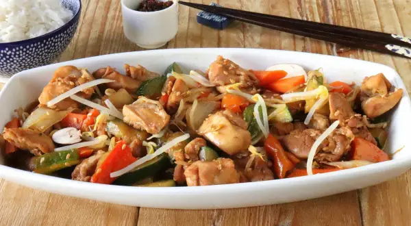 Un delicioso chop suey de pollo con verduras y brotes de soja, servido en un plato blanco junto a un tazón de arroz blanco y palillos.