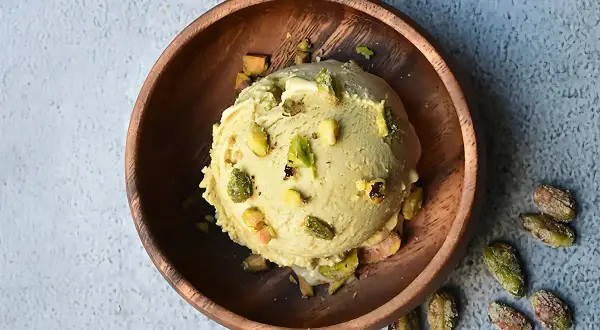 Cremoso helado de pistacho servido en un cuenco de madera, adornado con trozos de pistacho y acompañado de más pistachos enteros esparcidos.