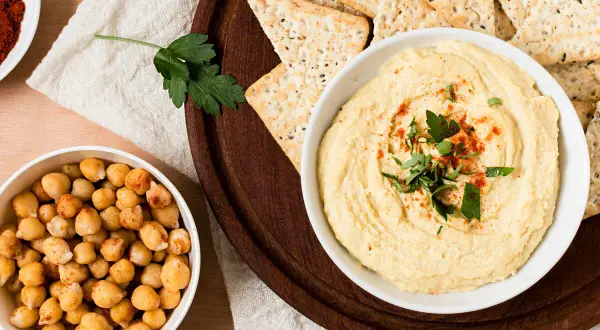 Un tazón de hummus cremoso, decorado con perejil y pimentón, junto a garbanzos y galletas saladas. ¡Perfecto para tu hummus receta!