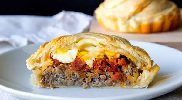 Un trozo de pastel de carne murciano, relleno de carne picada, tomate y huevo duro, envuelto en hojaldre dorado.
