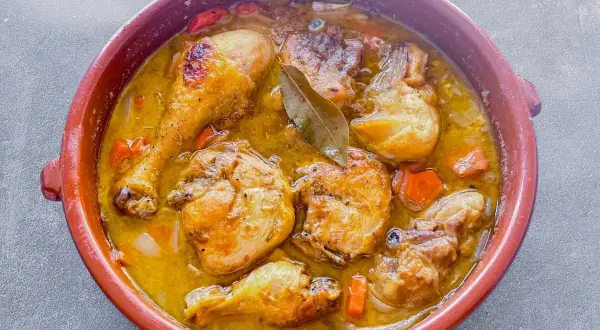 Sabroso pollo en salsa con trozos dorados, zanahorias y laurel en una cazuela de barro.