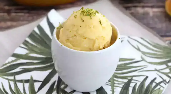 Una bola de sorbete de mango, refrescante y deliciosa, servida en una taza blanca sobre una servilleta con hojas tropicales.
