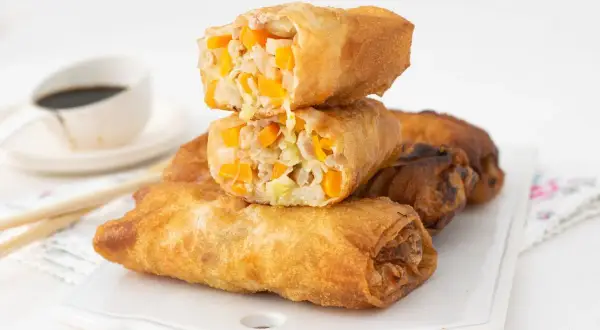 Rollitos de primavera dorados y crujientes, rellenos de verduras y carne. Se apilan sobre una bandeja blanca, con salsa de soja al fondo.