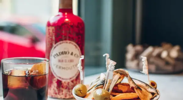 Un vaso de vermut con hielo y naranja, junto a una botella roja de Padró & Co. Vermouth y un plato de patatas y anchoas. ¡Perfecto para clases de vermut!