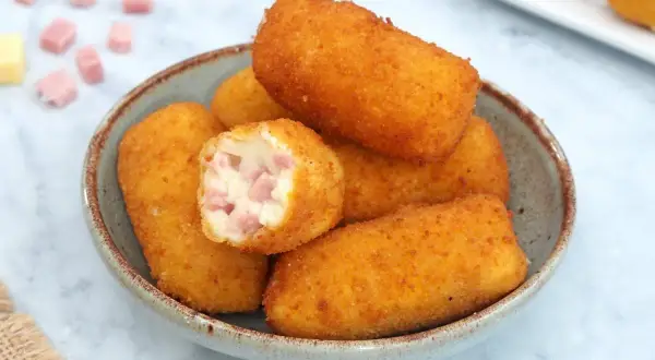 Un tazón lleno de crujientes croquetas de jamón, una de ellas partida mostrando su cremoso interior con trocitos de jamón.