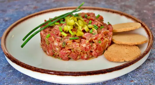 Tartar de salchichón picado finamente, adornado con cebollino y pepinillos, servido con tostas.