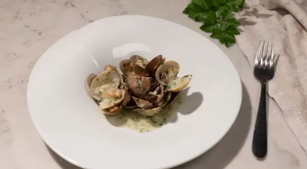 Un plato blanco con almejas en salsa verde, adornado con perejil fresco. Un tenedor y una servilleta completan la escena.