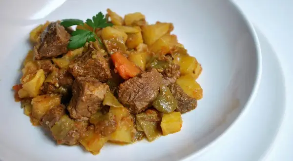 Un plato de carne guisada con patatas, zanahorias y pimientos verdes, adornado con perejil fresco.