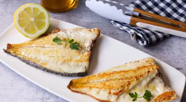 Dos filetes de dorada a la plancha, servidos en plato blanco con limón, perejil y utensilios de cocina.