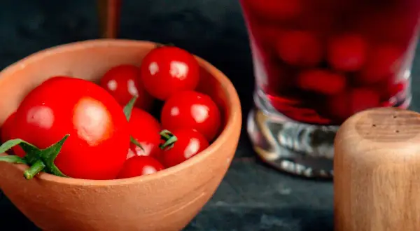 Un cuenco de barro lleno de tomates rojos brillantes, listos para un delicioso tartar de tomate. A su lado, un vaso con bebida roja y un salero de madera.