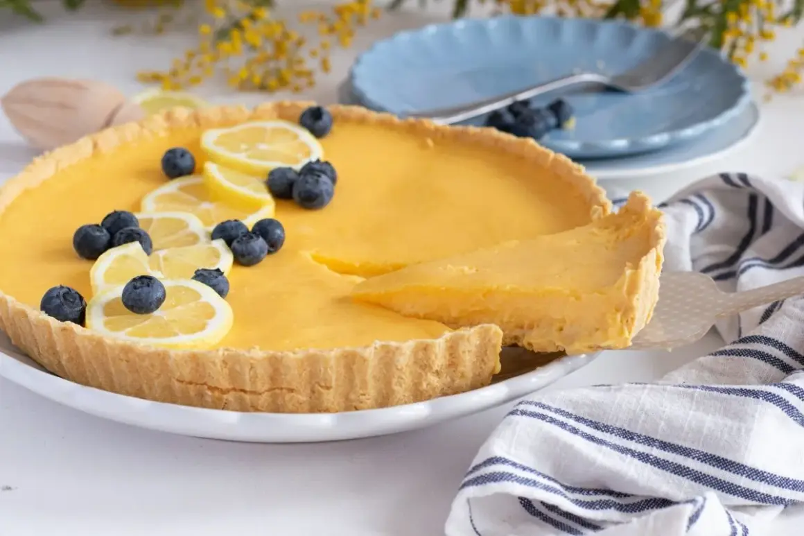Deliciosa tarta de limón, decorada con rodajas de limón y arándanos. Una porción se sirve con una espátula.