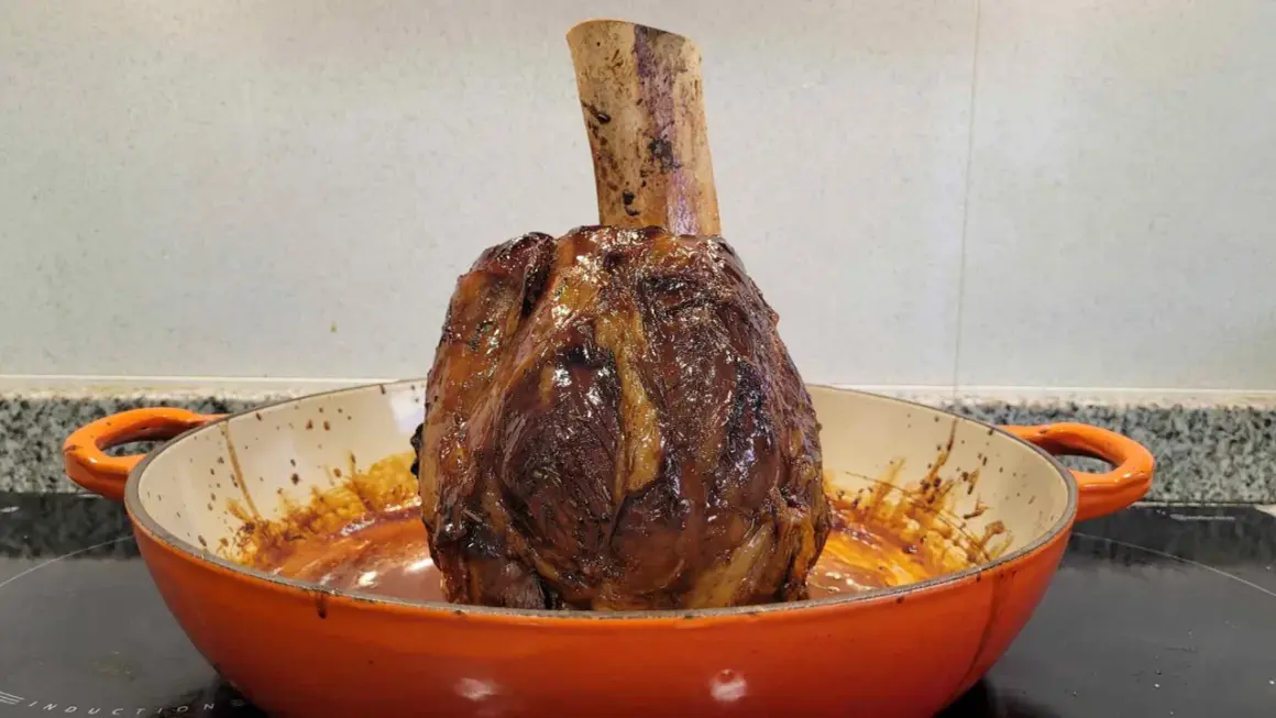 Un jugoso jarrete de ternera asado, con su hueso expuesto, reposa en una cazuela naranja, listo para ser disfrutado.