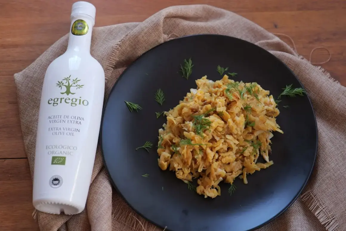 Un plato de revuelto de bacalao con eneldo fresco, acompañado de una botella de aceite de oliva virgen extra ecológico Egregio.