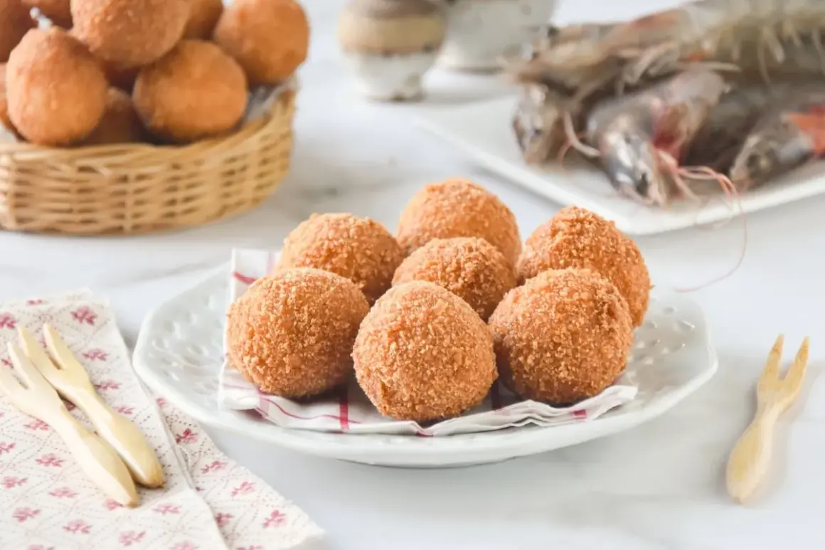 Croquetas de gambas doradas y crujientes en un plato blanco, con más en una cesta de mimbre al fondo.