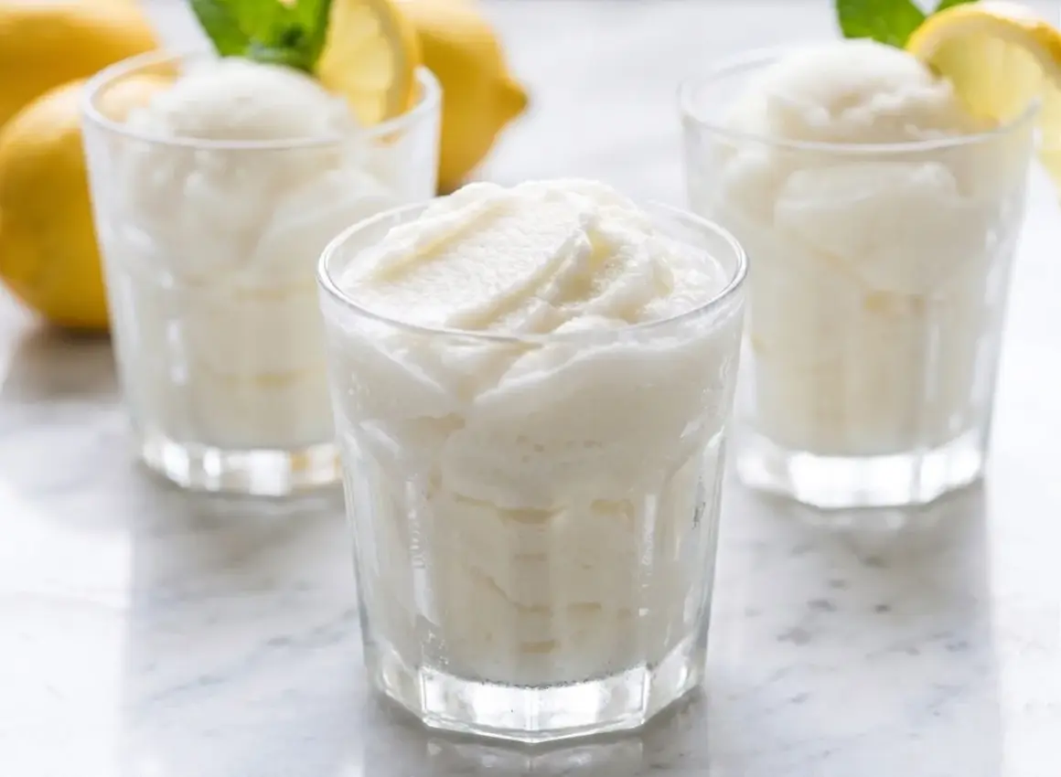 Tres vasos de sorbete de limón refrescante, con limones amarillos y hojas de menta en el fondo.