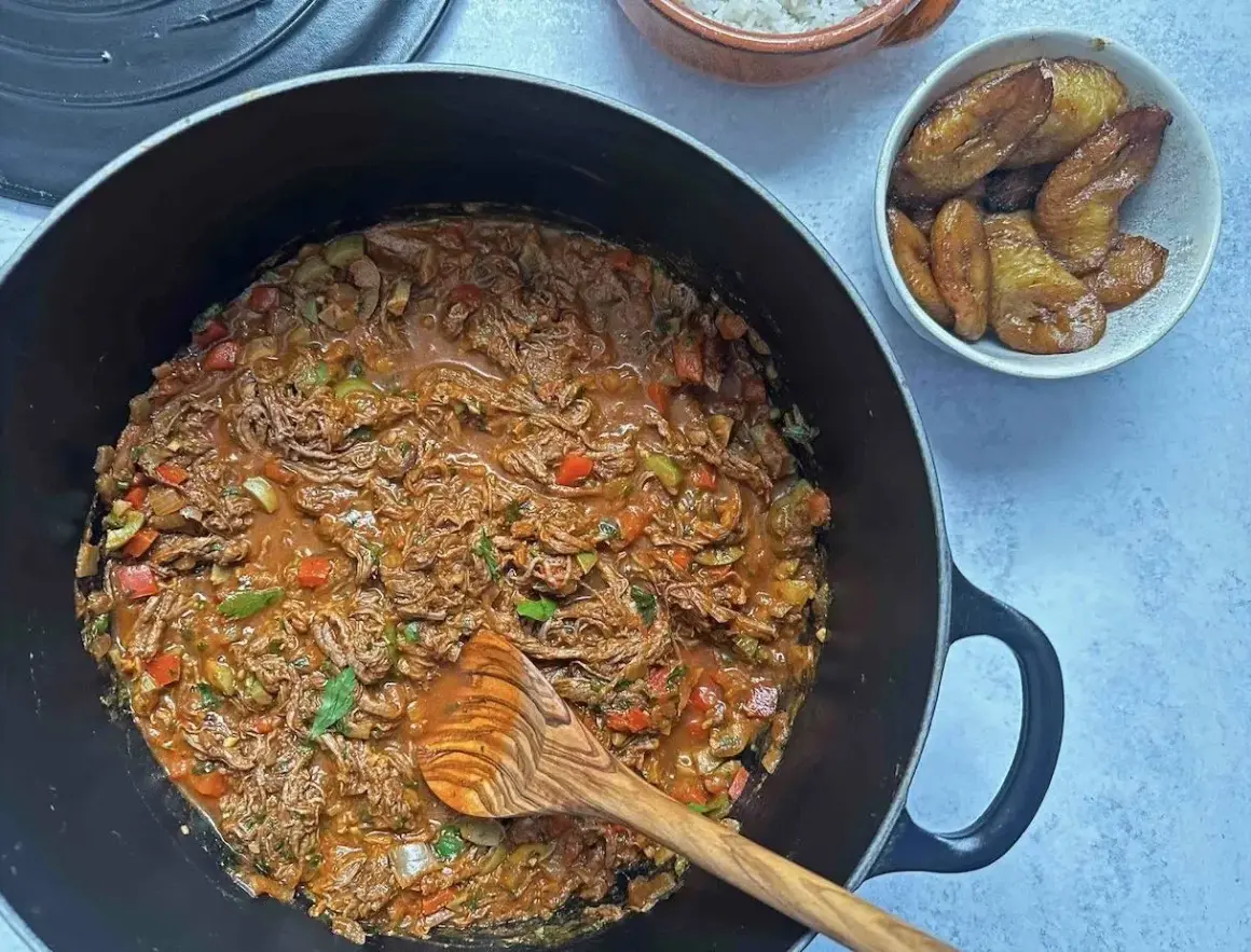 Un delicioso plato de ropa vieja, con trozos de carne deshebrada en salsa roja, acompañado de plátanos maduros fritos y arroz blanco.