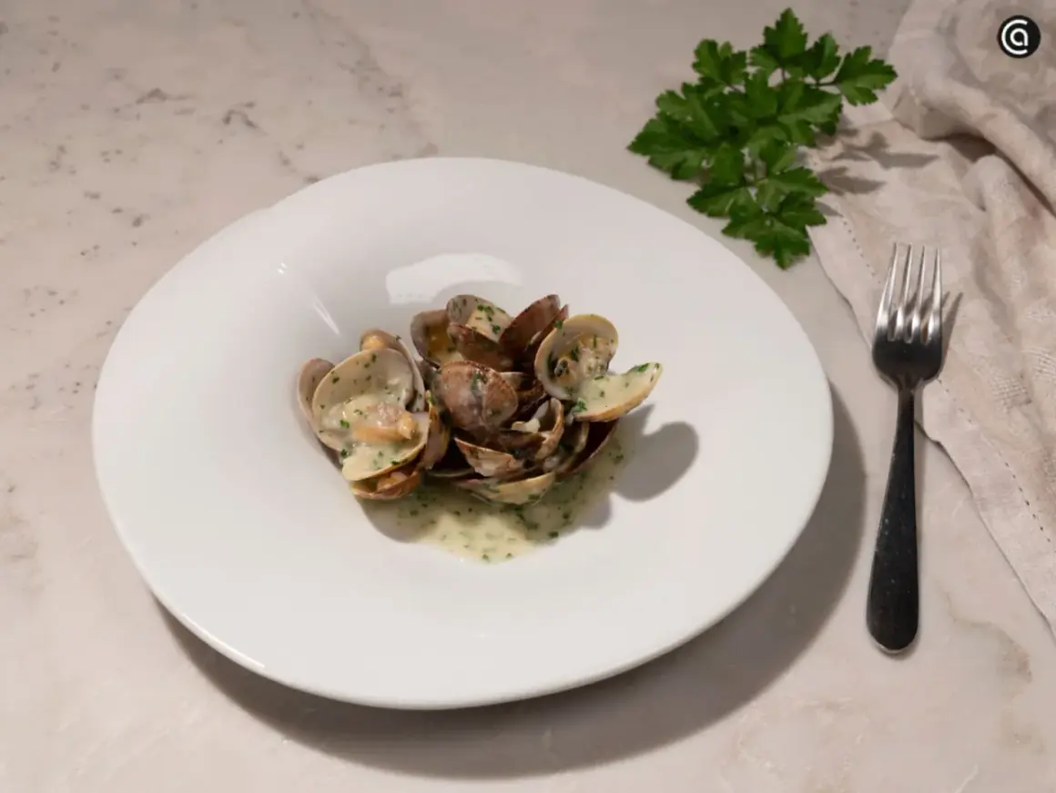 Un plato blanco con almejas en salsa verde, adornado con perejil fresco. Un tenedor y una servilleta completan la escena.