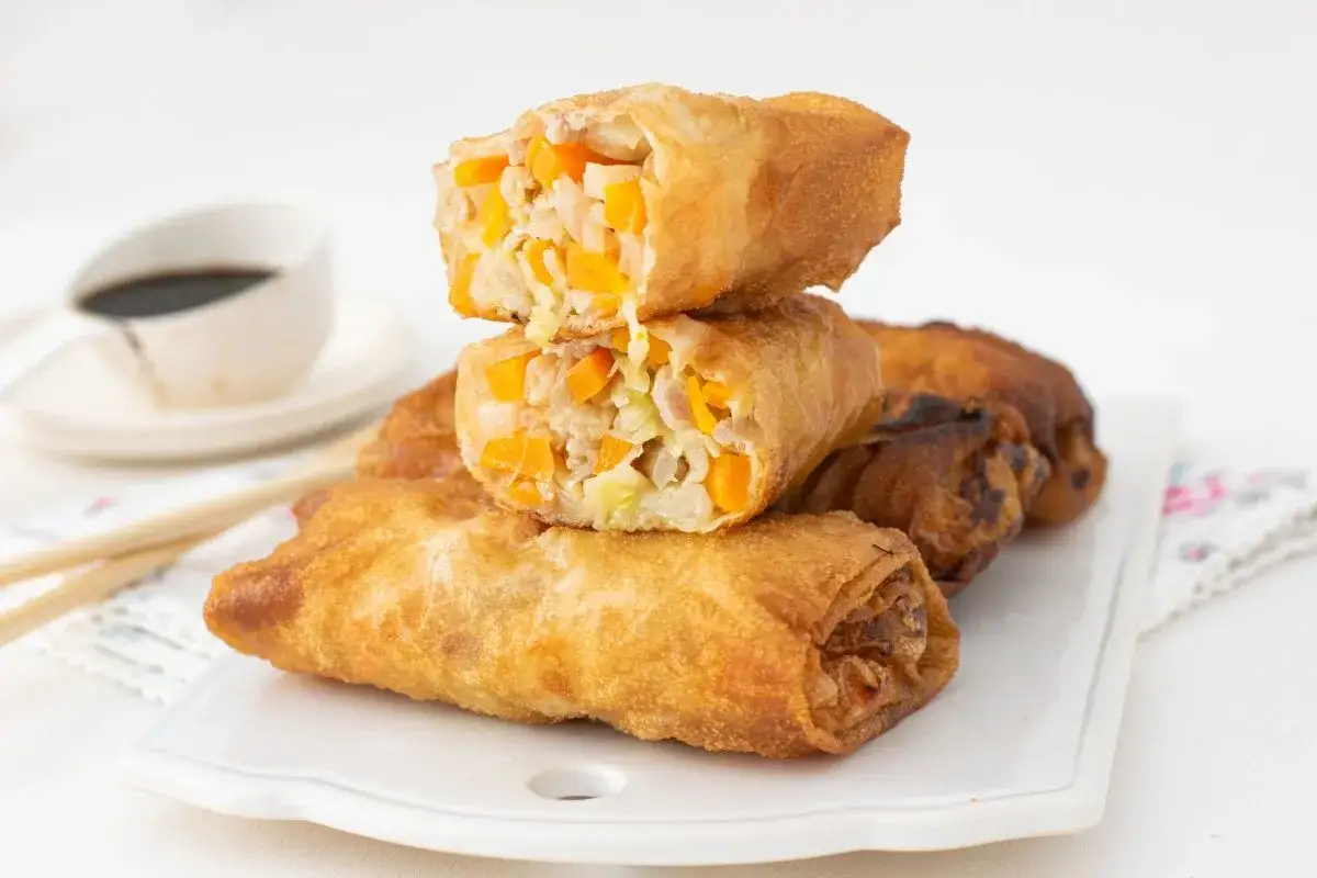 Rollitos de primavera dorados y crujientes, rellenos de verduras y carne. Se apilan sobre una bandeja blanca, con salsa de soja al fondo.