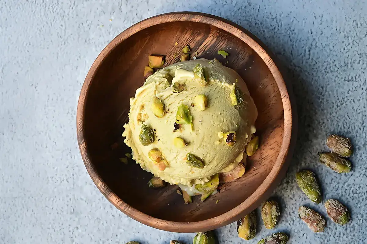 Cremoso helado de pistacho servido en un cuenco de madera, adornado con trozos de pistacho y acompa&ntilde;ado de m&aacute;s pistachos enteros esparcidos alrededor.