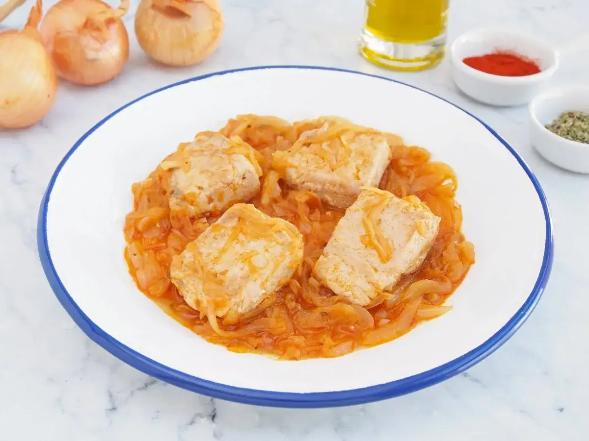 Un bonito encebollado servido en un plato blanco con borde azul. El pescado está cubierto de cebolla caramelizada y salsa.