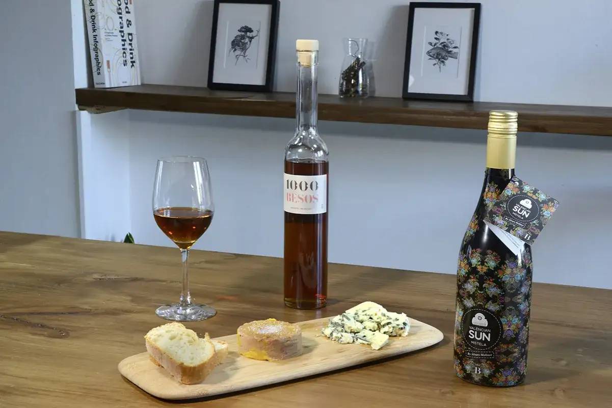 Una copa de vino dulce, pan, paté y queso azul acompañan a dos botellas de mistela, una de ellas decorada con flores.