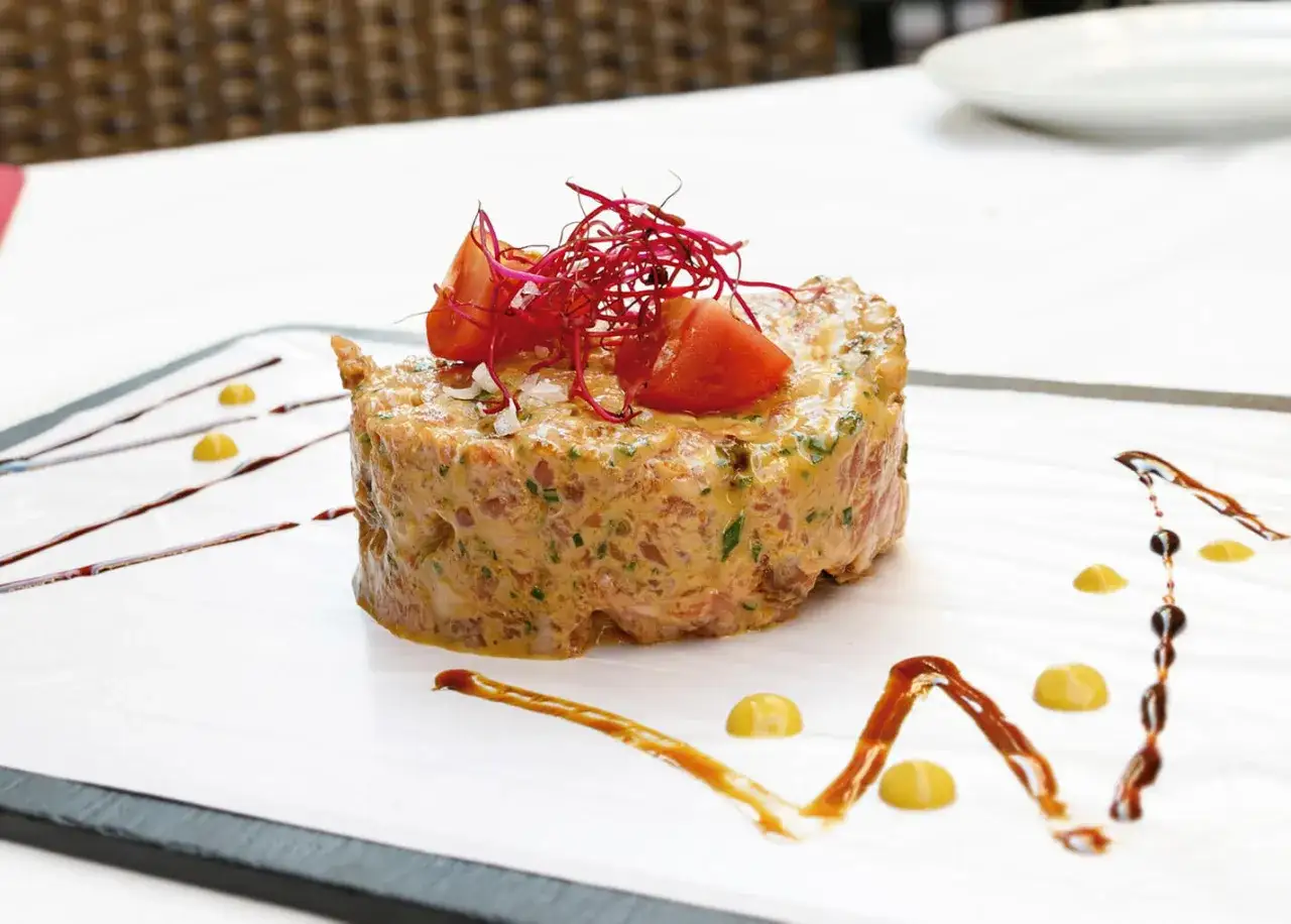 Delicioso tartar de salchich&oacute;n, adornado con tomate y brotes rojos, servido en un plato blanco con salsas decorativas.