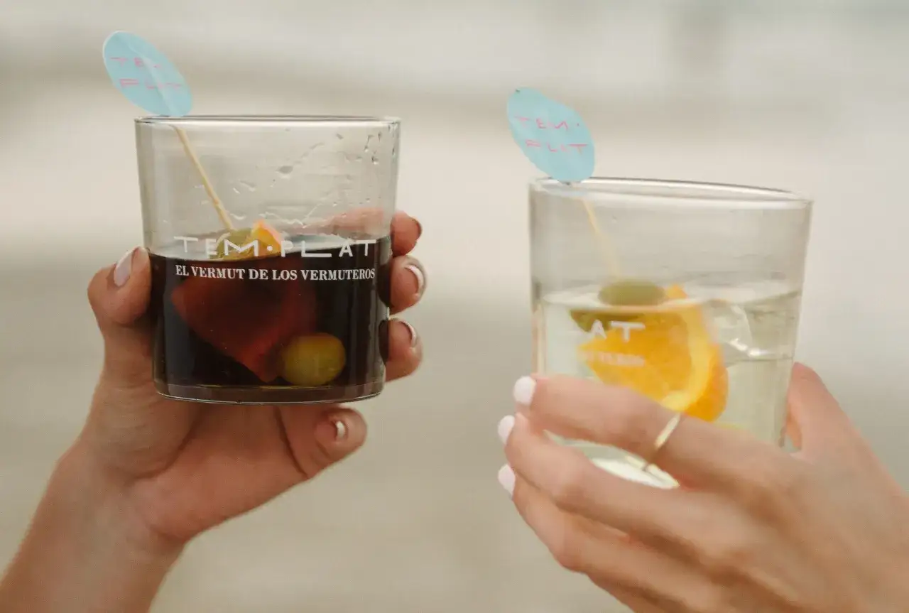 Dos manos sostienen vasos con vermut. Uno es rojo con aceituna y naranja, el otro blanco con naranja. Ambos tienen el nombre