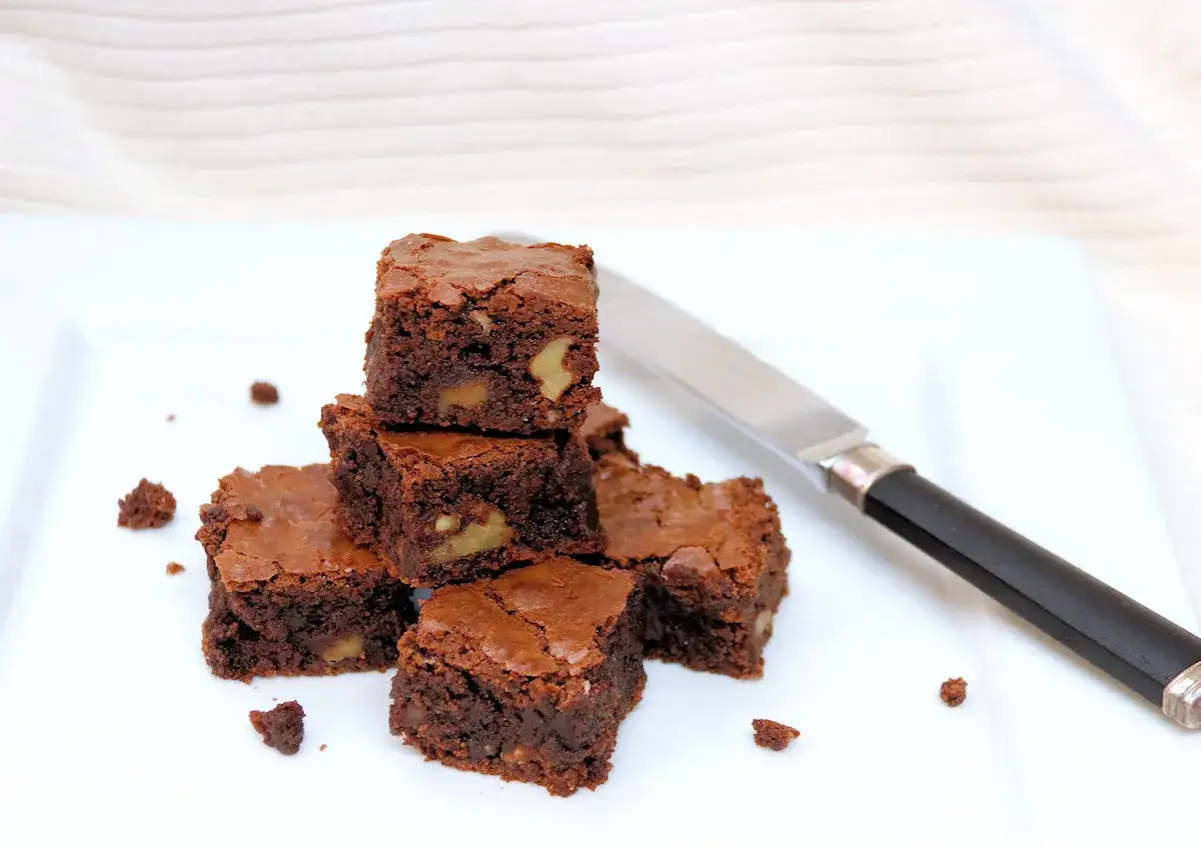 Deliciosos trozos de brownie sin gluten con nueces, apilados en un plato blanco junto a un cuchillo.