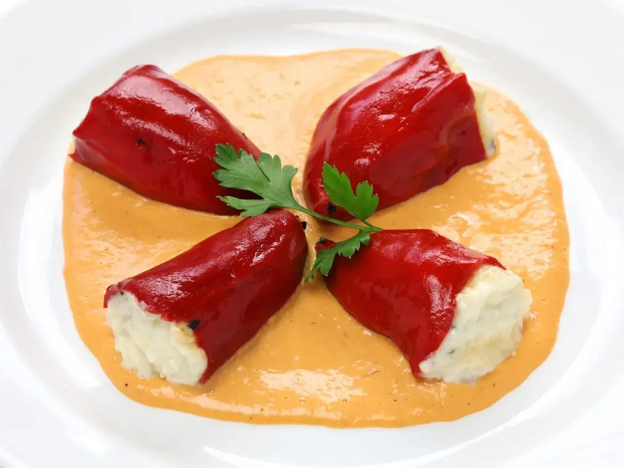 Cuatro pimientos rojos rellenos de bacalao, servidos sobre una cremosa salsa naranja y decorados con perejil.