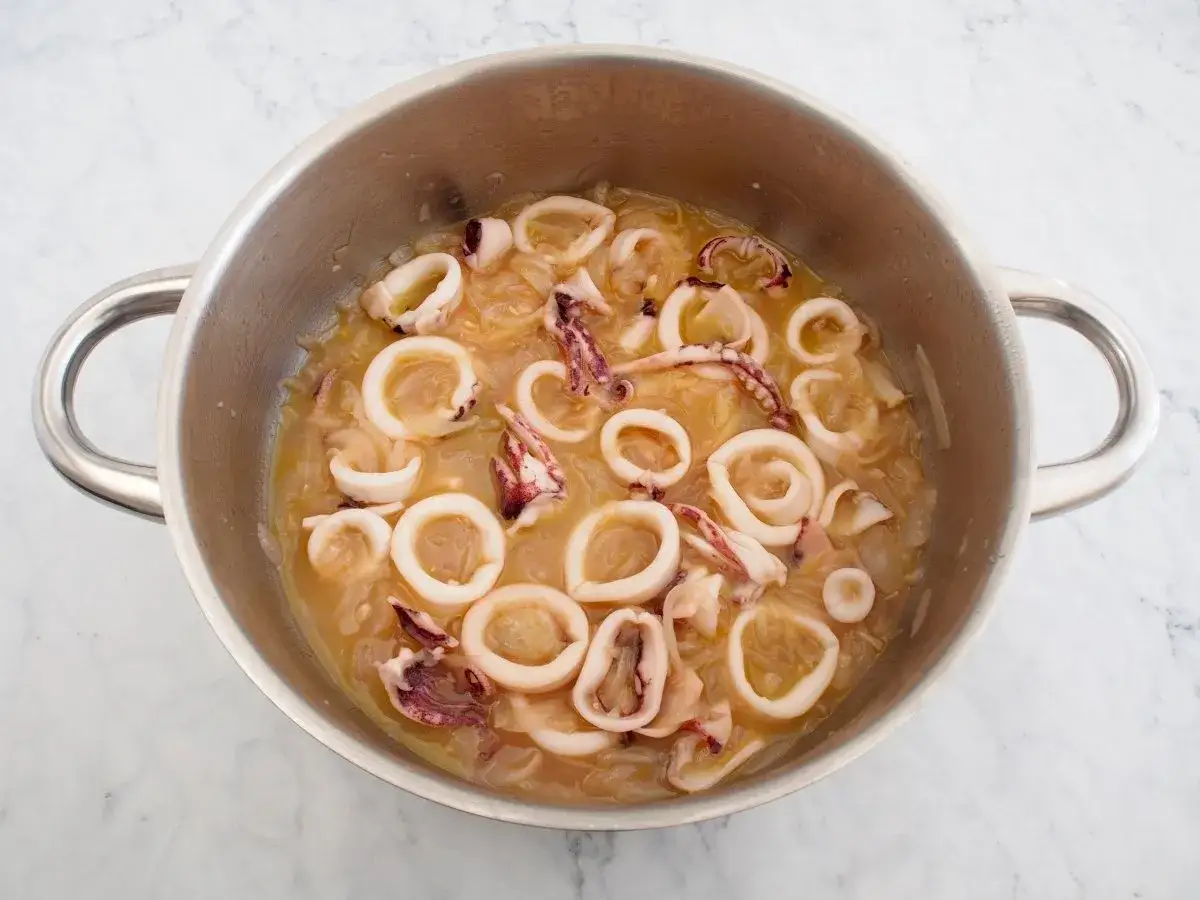 Calamares encebollados en una olla de metal, con aros de calamar y tentáculos cocinándose en una salsa dorada con cebolla caramelizada.