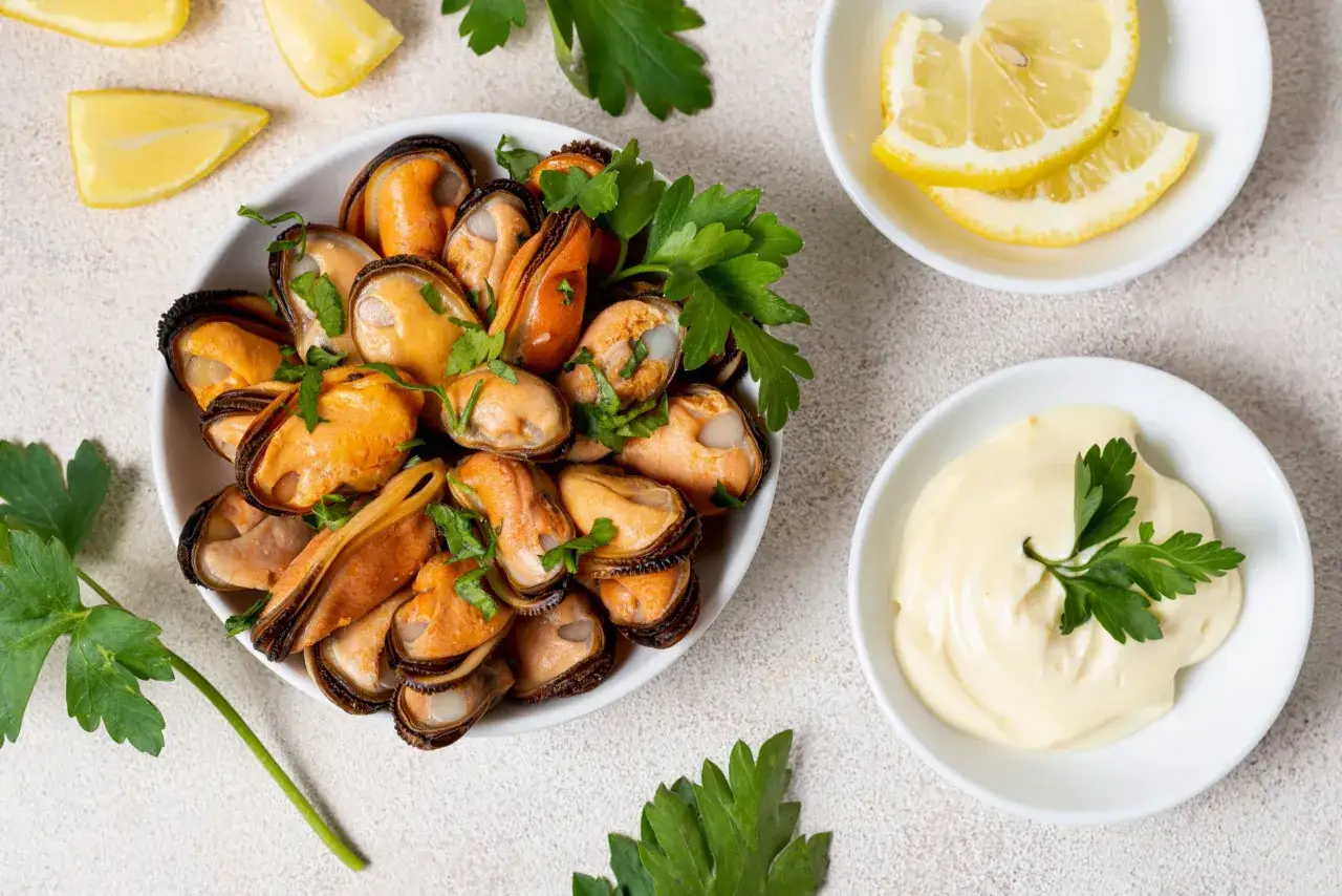 Un plato de mejillones al vapor, decorado con perejil fresco, acompañado de rodajas de limón y una salsa cremosa.