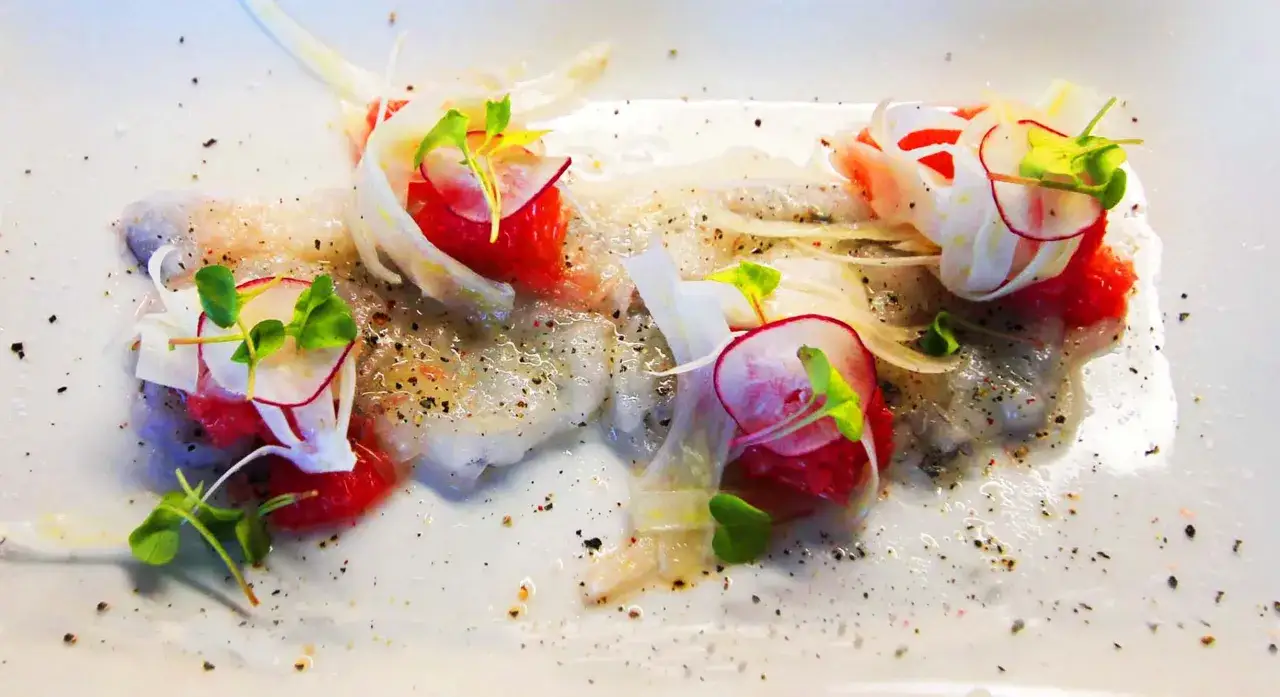 Delicado carpaccio de gambas con finas láminas de rábano, cebolla y brotes verdes, aderezado con pimienta negra.