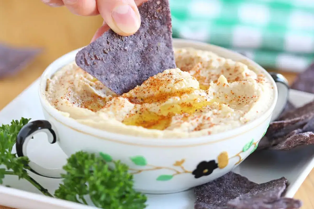 Un chip azul se sumerge en cremoso hummus sin tahini, espolvoreado con piment&oacute;n y aceite de oliva.