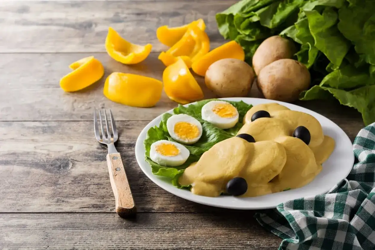 Papa a la huanca&iacute;na servida en un plato blanco con lechuga, huevos duros y aceitunas negras. Al fondo, pimientos amarillos y papas.