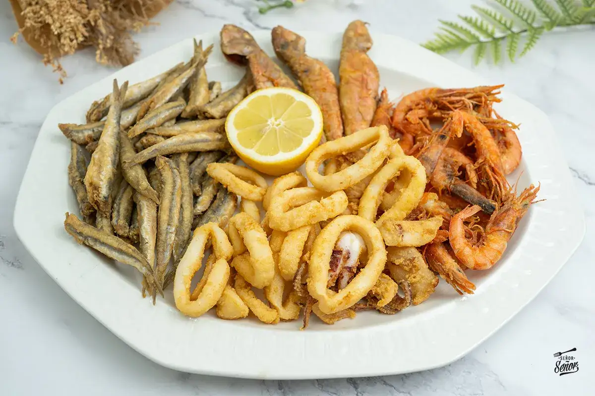 Plato de fritura de pescado con boquerones, calamares, gambas y trozos de pescado rojo, adornado con una rodaja de lim&oacute;n.