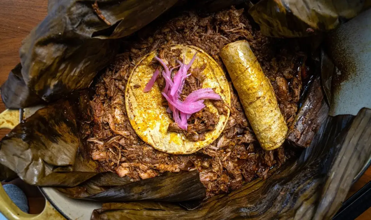 Cochinita pibil jugosa envuelta en hoja de plátano, servida con tortilla y cebolla morada encurtida.