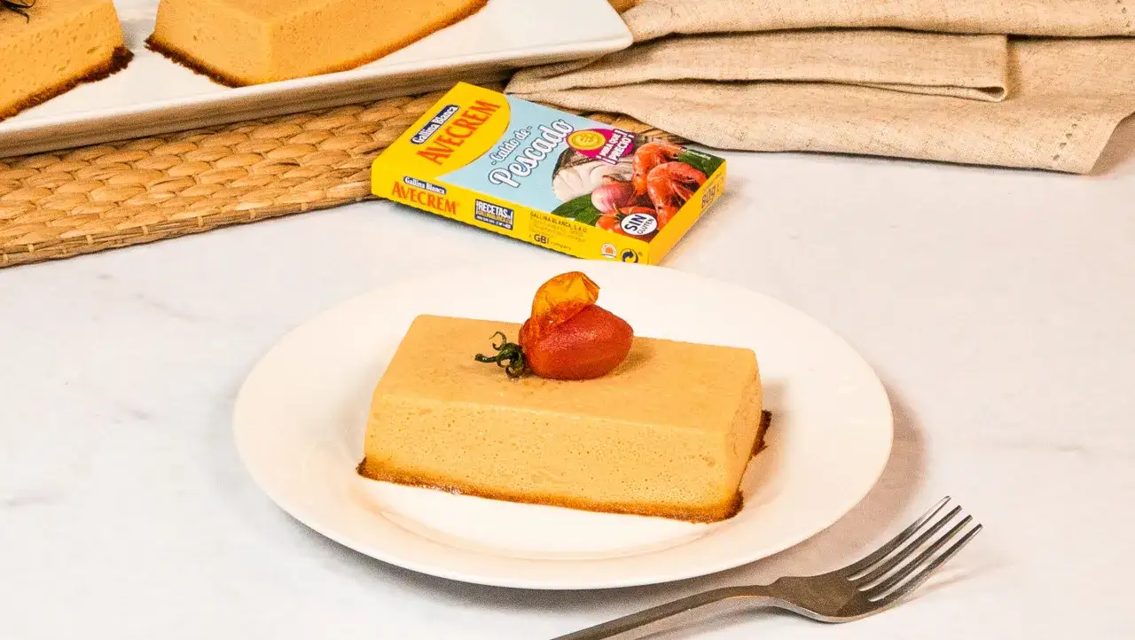 Un trozo de pastel de pescado, decorado con un tomate cherry y una hoja verde, servido en un plato blanco junto a un tenedor.
