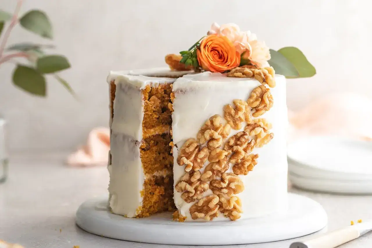 Pastel de zanahoria cubierto de glaseado blanco, decorado con nueces y flores naranjas.