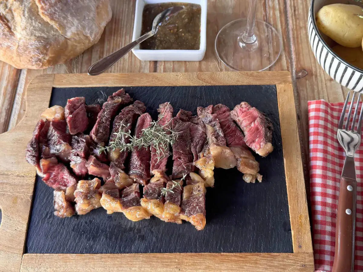 Un jugoso entrecot de ternera cortado en rodajas, adornado con romero fresco, servido sobre pizarra. Acompa&ntilde;ado de pan, salsa y patatas.