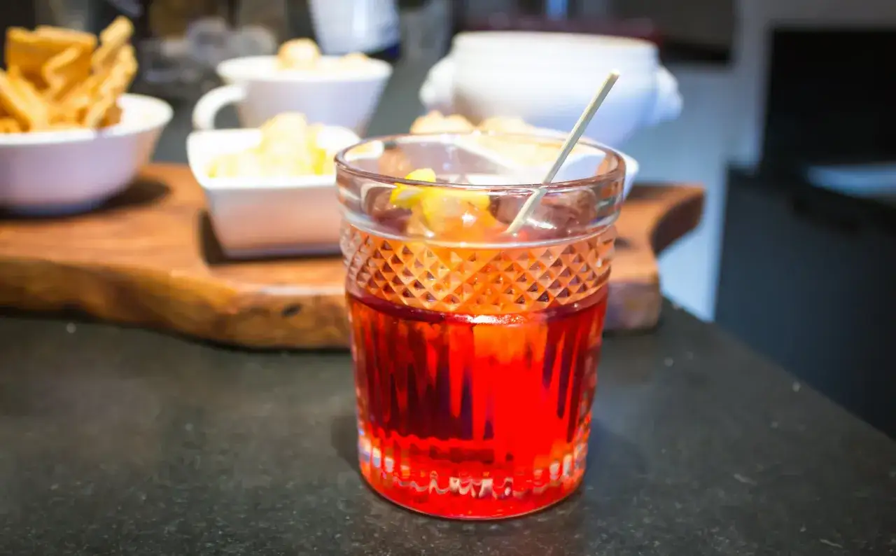 Un vaso de vermut rojo con hielo y una rodaja de lim&oacute;n, acompa&ntilde;ado de aperitivos.