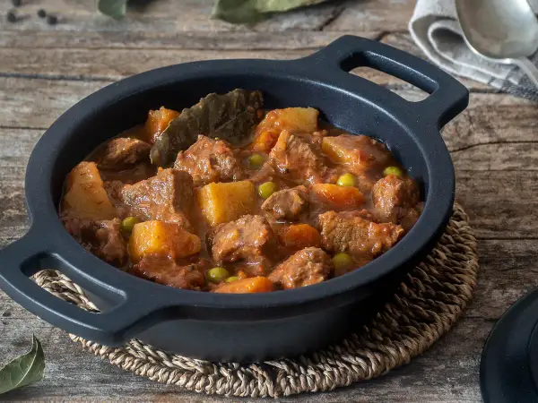 Estofado de ternera con patatas, guisantes y laurel, servido en cazuela negra sobre mesa de madera.