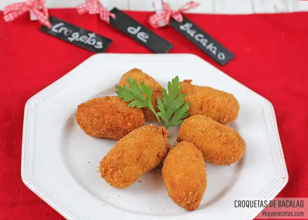 Seis croquetas de bacalao doradas y crujientes, adornadas con perejil fresco, servidas en un plato blanco.