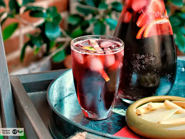 Un refrescante tinto de verano con hielo y rodajas de limón en un vaso, junto a una jarra llena de la misma bebida y un cuenco con palitos de queso.