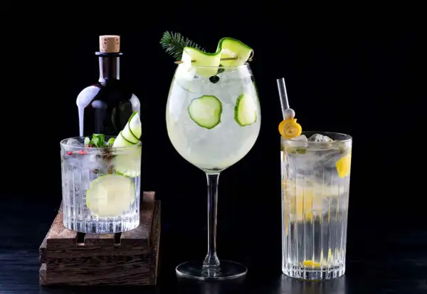 Tres refrescantes cócteles de ginebra, decorados con pepino y limón, junto a una botella de ginebra.