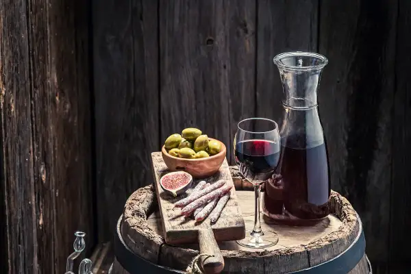 Copa de vino tinto, aceitunas, higo y salchichones sobre una tabla de madera, junto a una jarra de vino. Un festín que evoca la pregunta: ¿qué es el moscatel?