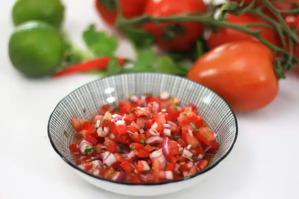Un tazón de pico de gallo fresco, con tomates, cebolla morada y cilantro picados. Al fondo, tomates, limas y chiles rojos.