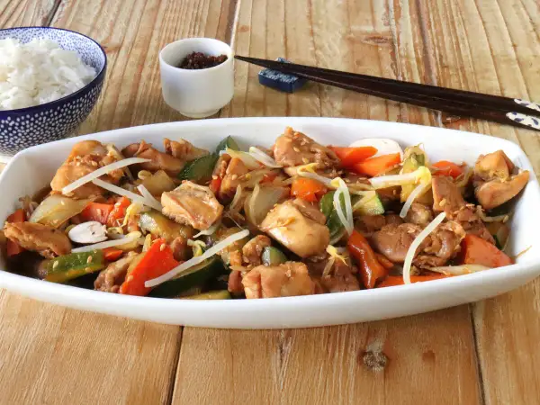 Un delicioso chop suey de pollo con verduras y brotes de soja, servido en un plato blanco junto a un tazón de arroz blanco y palillos.